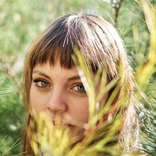 Mulher com olhos verdes e cabelo castanho com madeixas, rodeada por vegetação natural, com luz suave e tons dourados que evocam as tendências de beleza de outono.